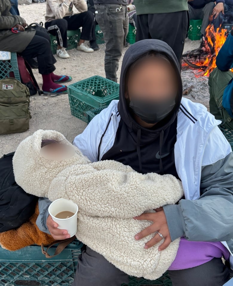 A woman and her child sit outside, beside a fire. Their faces have been blurred.