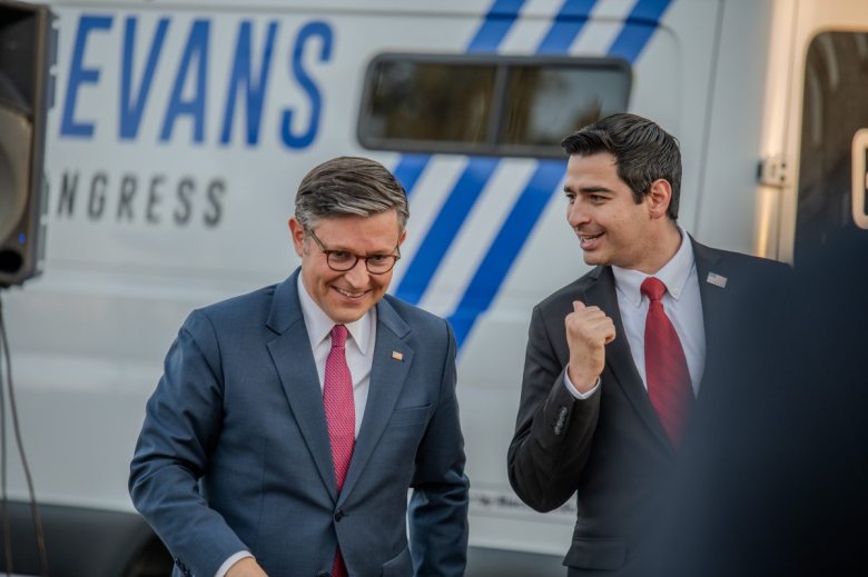 Two men in suits and red ties stand in front of a vehicle with campaign text. One man has glasses and is smiling, while the other appears to be speaking energetically.