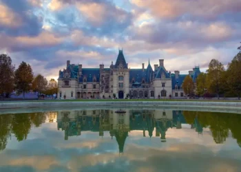 A large, ornate castle with multiple spires and turrets set against a cloudy sky, reflected in a tranquil pond in the foreground. Trees surround the expansive property.