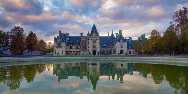 A large, ornate castle with multiple spires and turrets set against a cloudy sky, reflected in a tranquil pond in the foreground. Trees surround the expansive property.