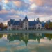 A large, ornate castle with multiple spires and turrets set against a cloudy sky, reflected in a tranquil pond in the foreground. Trees surround the expansive property.