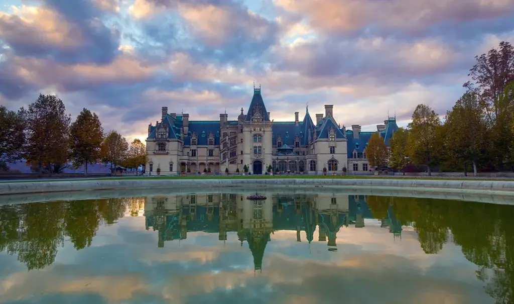 A large, ornate castle with multiple spires and turrets set against a cloudy sky, reflected in a tranquil pond in the foreground. Trees surround the expansive property.