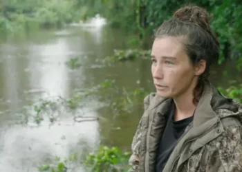 A woman in a green jacket stood in front of a flooded road. She is to the right of the frame and had black hair tied up.