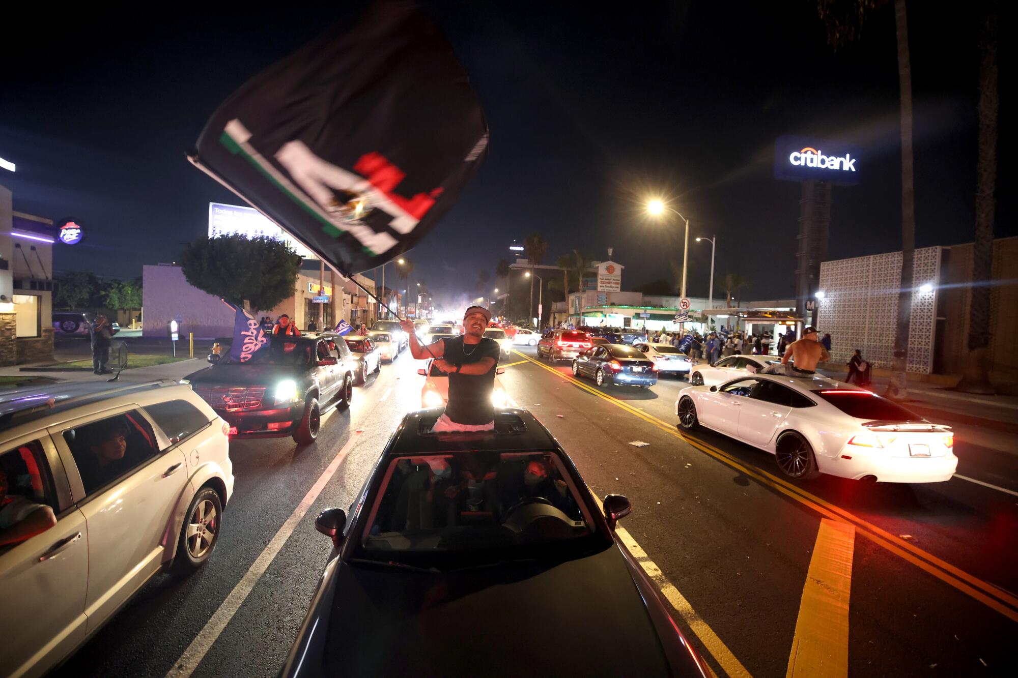 A man from an open sunroof of a car waves a Dodgers flag at night 