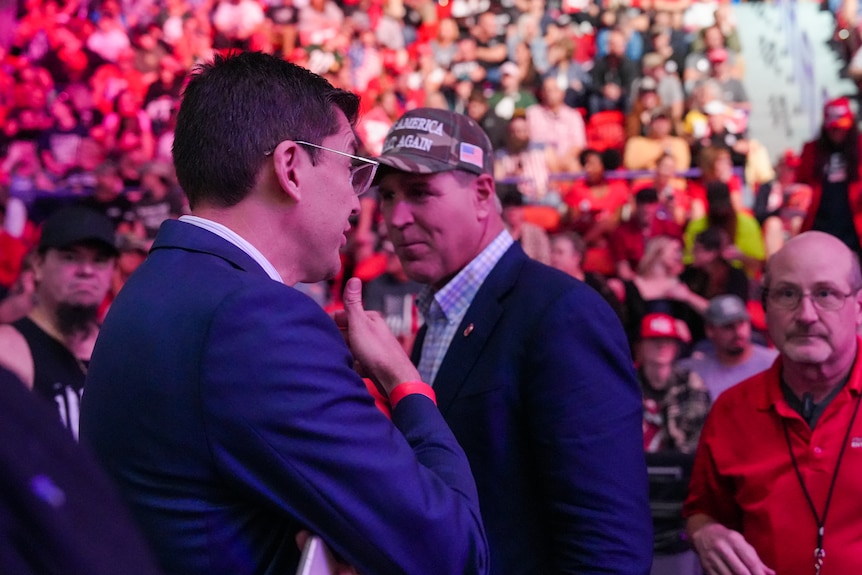 A man in a MAGA hat and suit jacket chat to another man in a suit jacket 