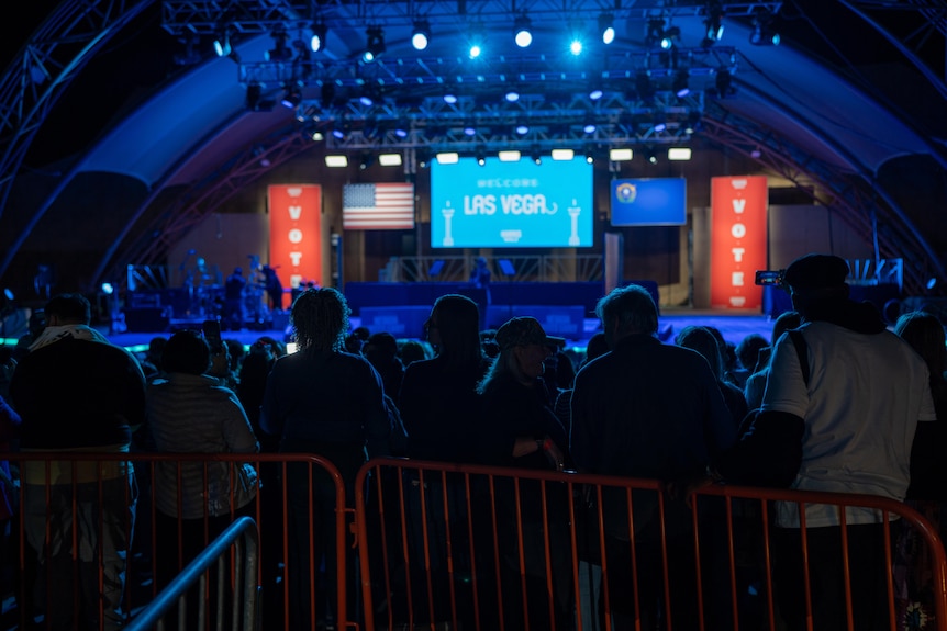 a crowd waits in front of an empty stage with a US flag and a screen that reads Las Vegas