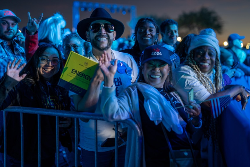 a woman in a Harris hat waves to the camera in a happy-looking crowd