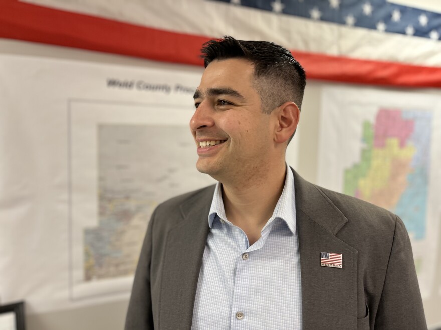 A man wearing a gray suit jacket with an American flag pin on his lapel stands smiling