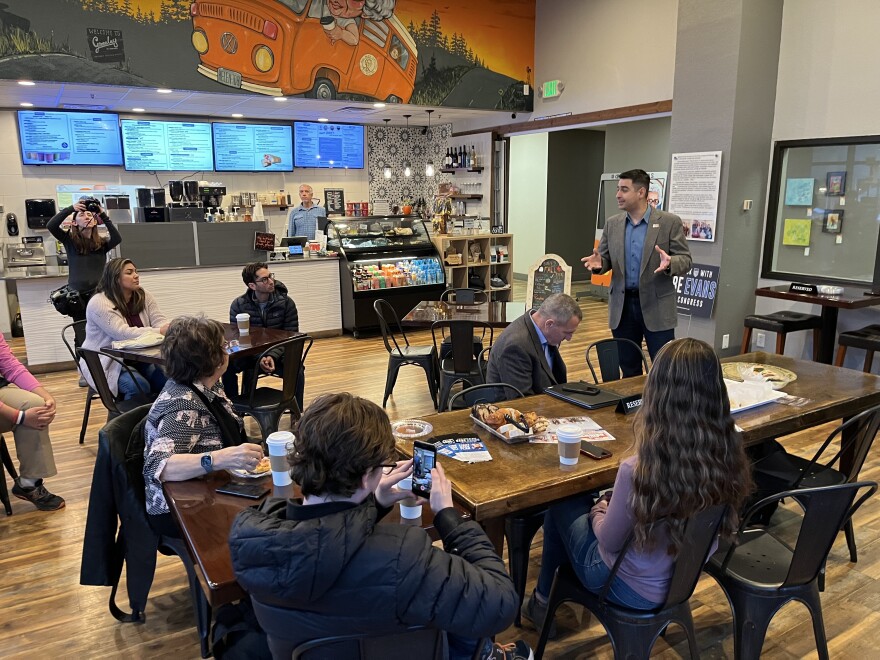 Aman in a gray jacket and blue shirt stands in a restaurant speaking while those seated at tables listen. 