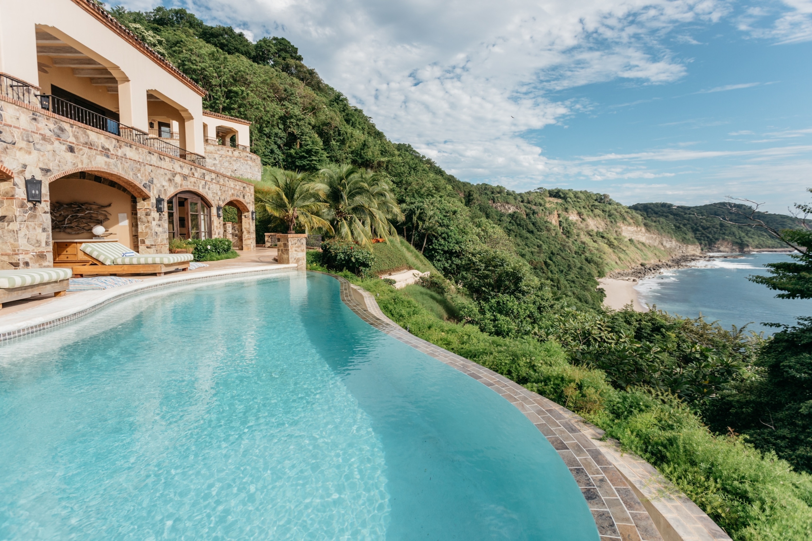 A two-storied stone building with terraces and a turquoise blue pool overlooking a hill covered in green tropical foliage which leads down to the sea.