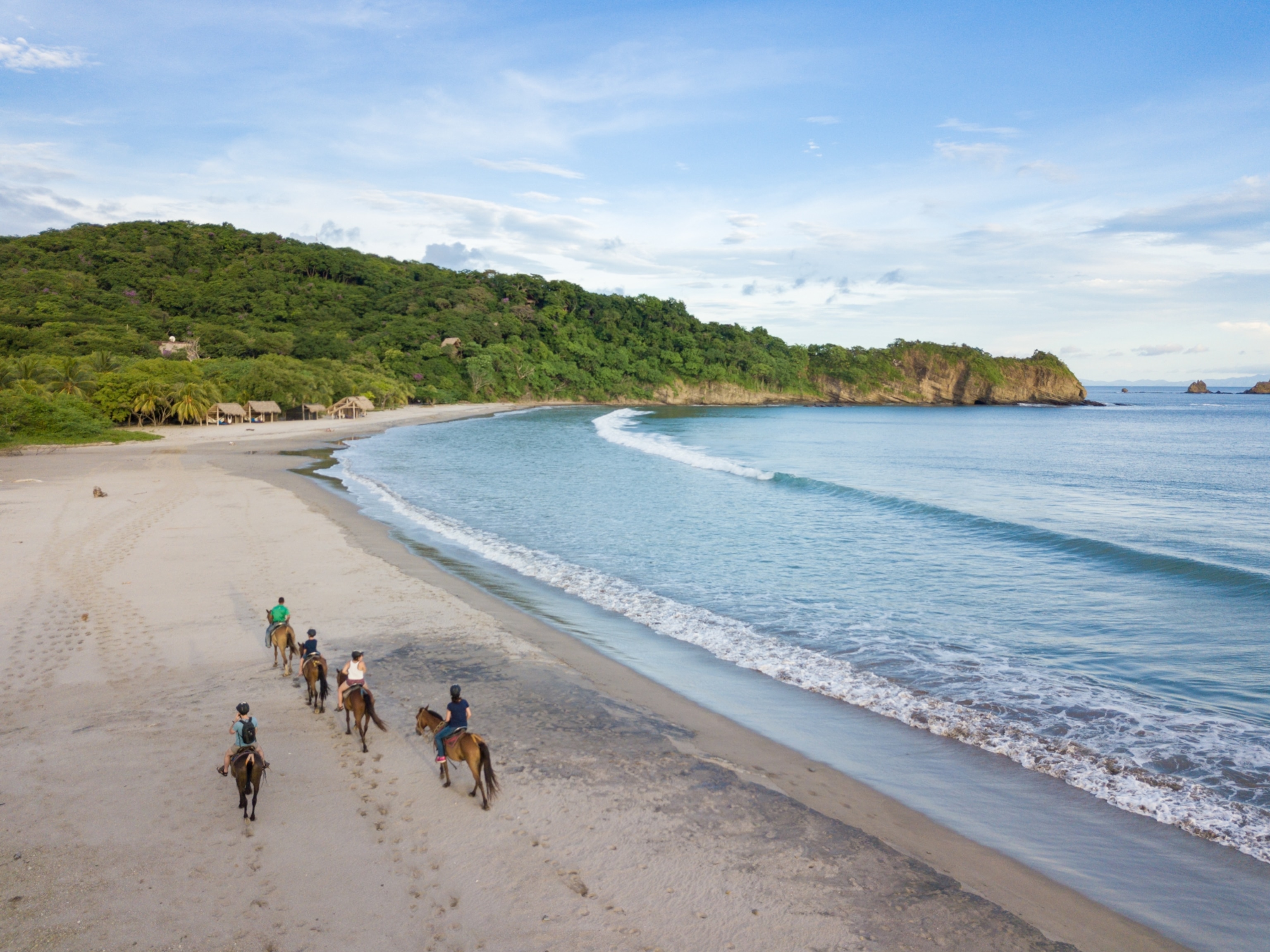 An elevated view of a group of tourists riding horseback along a beach with gentle waves. In the distance, bungalows are seen tucked amongst jungle foliage.