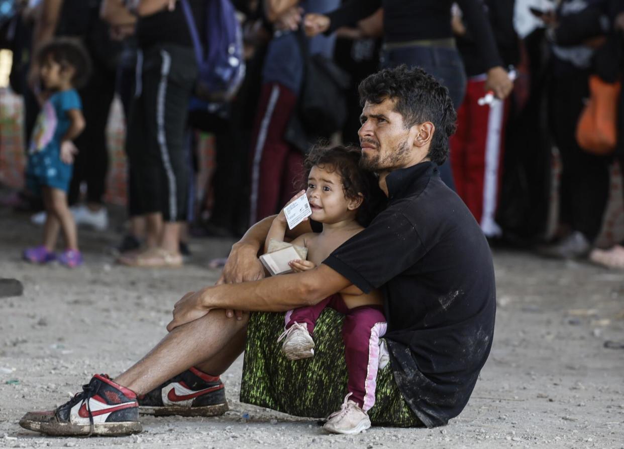 Migrants wait to load buses in a border patrol holding area, including a man and child sitting on the ground.