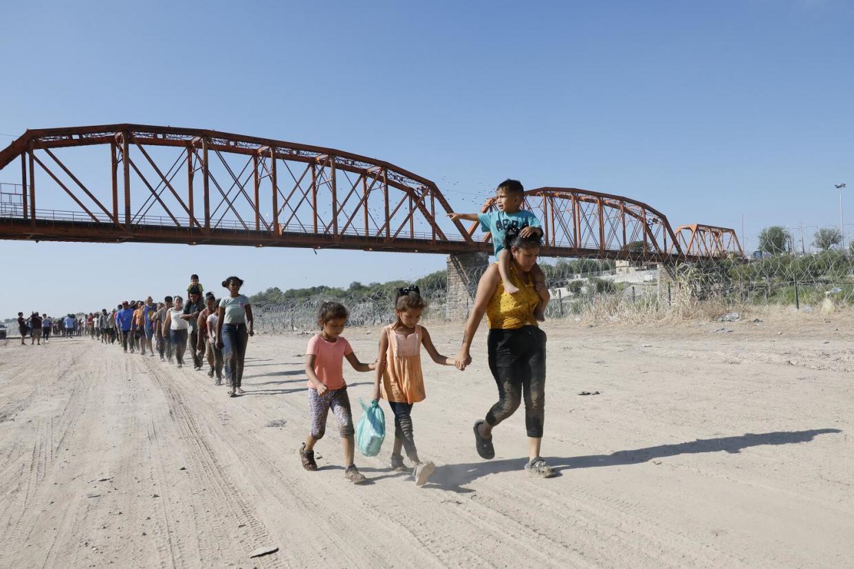 Hundreds of migrants walk under a bridge on a dirt path, including adults and children.