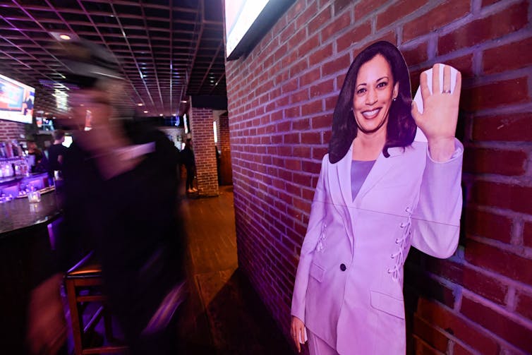 A cardboard cut out of a woman with brown hair and a pantsuit waves and stands against a brick wall, while blurred people are seen at a dimly lit bar.