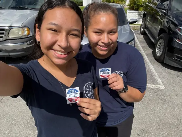 Christell and Leah take a selfie in their blue outfits, holding a sticker that says 