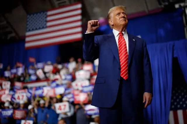 Donald Trump, dressed in a blue suit and red tie, raises a clenched fist in front of the American flag and his voters