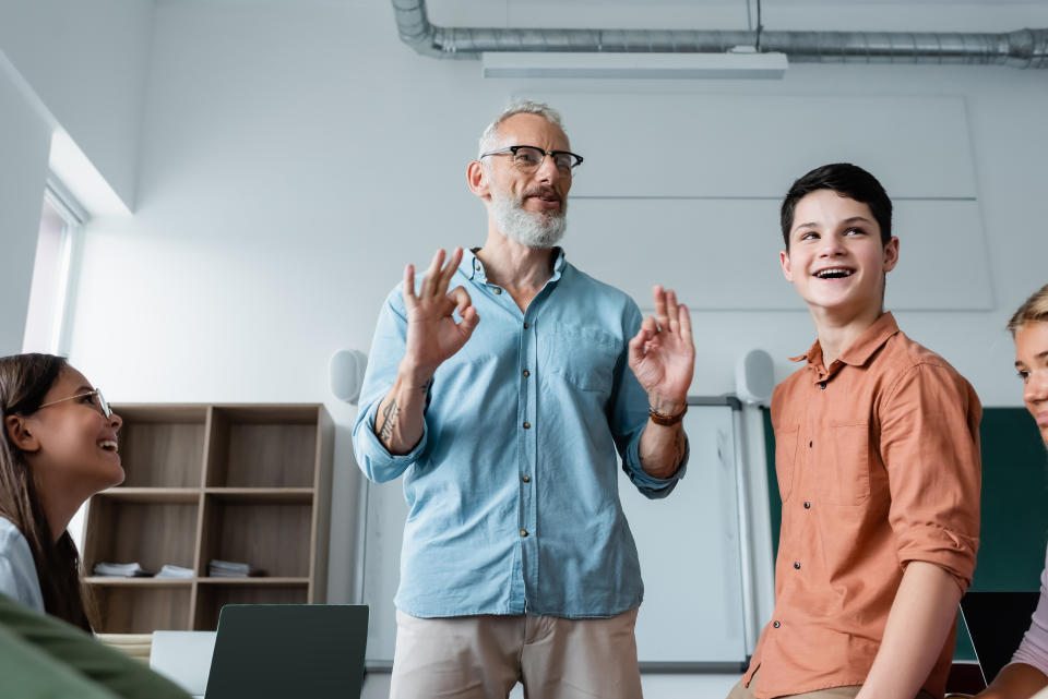 A teacher in a casual shirt engages with smiling students in a classroom discussion