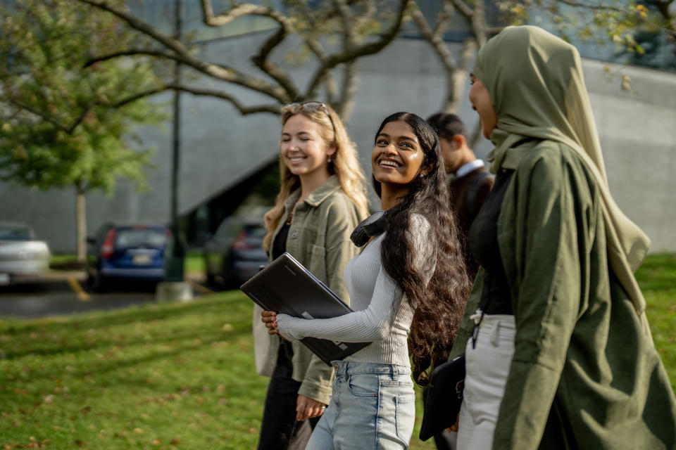 Three people walk outside, smiling and talking. One holds a laptop. Trees and parked cars are in the background