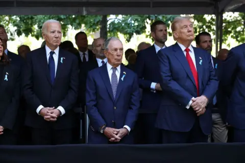 Getty Images Joe Biden, Michael Bloomberg and Donald Trump stand in a line on the front row of a memorial service to remember the victims of the 9/11 attacks. 

There are several people stood behind them, all three have their hands clasped together in front of them. 