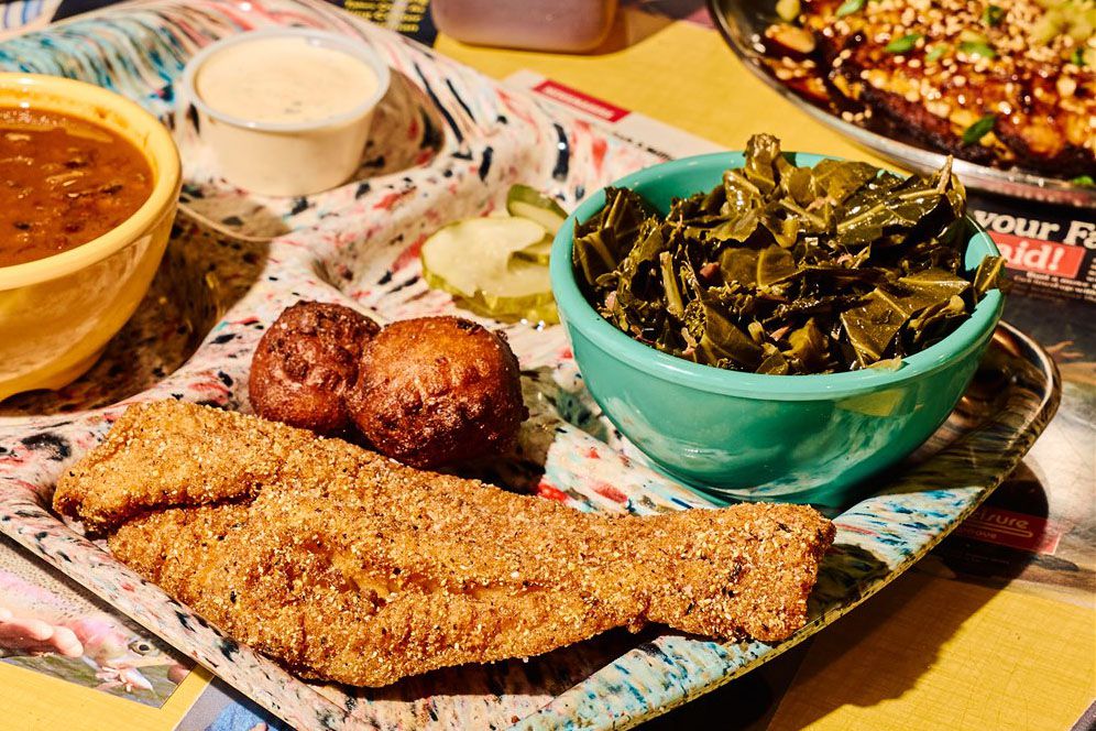 A tray holds a piece of fried fish, hush puppies, a bowl of collard greens, sliced pickles, a bowl of soup, and a container of white sauce.