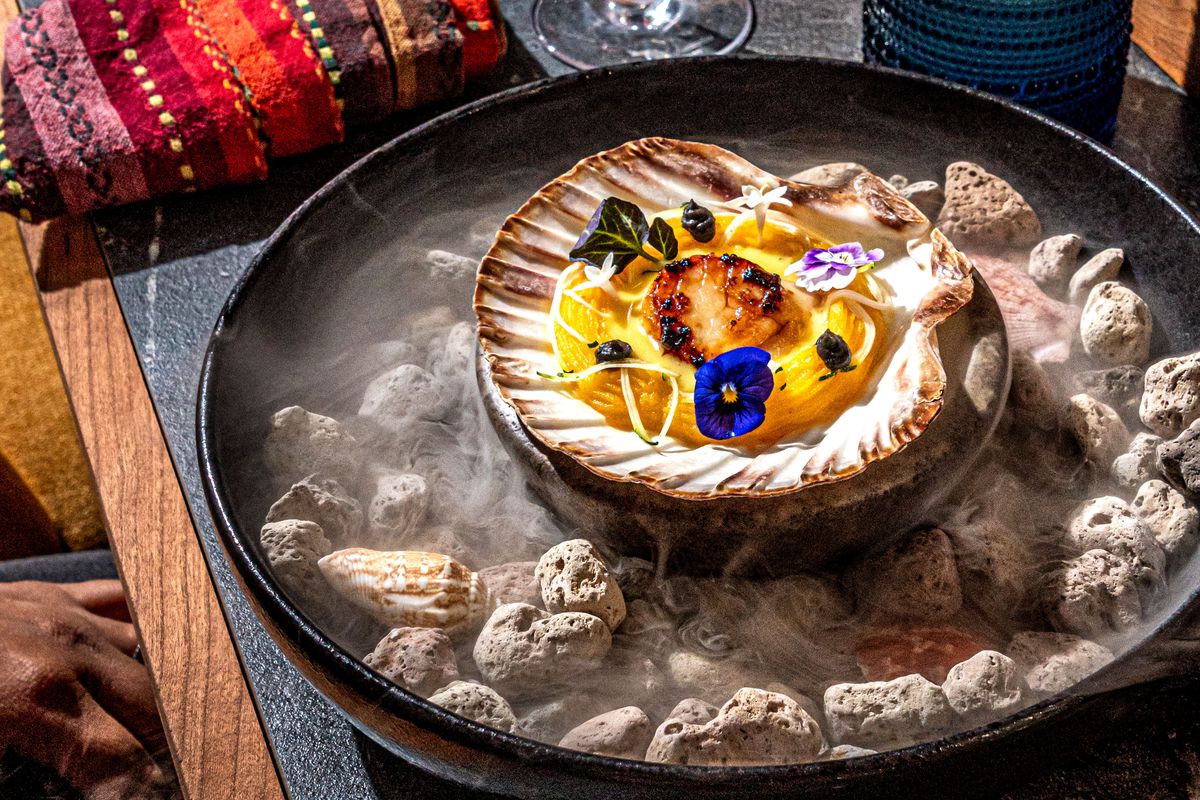 An image of the Three Sisters dish, which includes squash placed in a large scallop shell half, and topped with a scallop placed in the center, which is surrounded by royal blue and forest green flowers. The dish is sitting in a bowl with gray rocks and steam rising from the bowl.