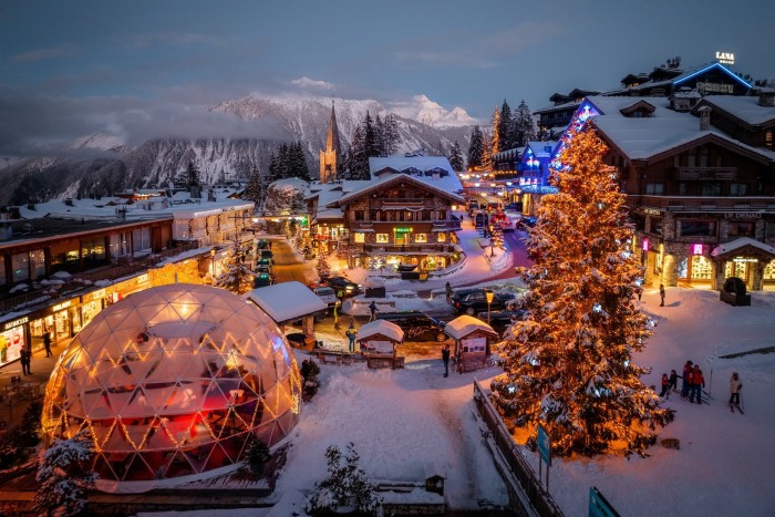 A snow-covered village at night. A pine tree in the foreground is flecked with snow and fairy lights