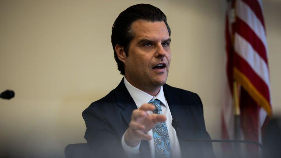 Matt Gaetz, wearing a suit, gestures with his first hand while sitting and speaking at a microphone during a hearing 