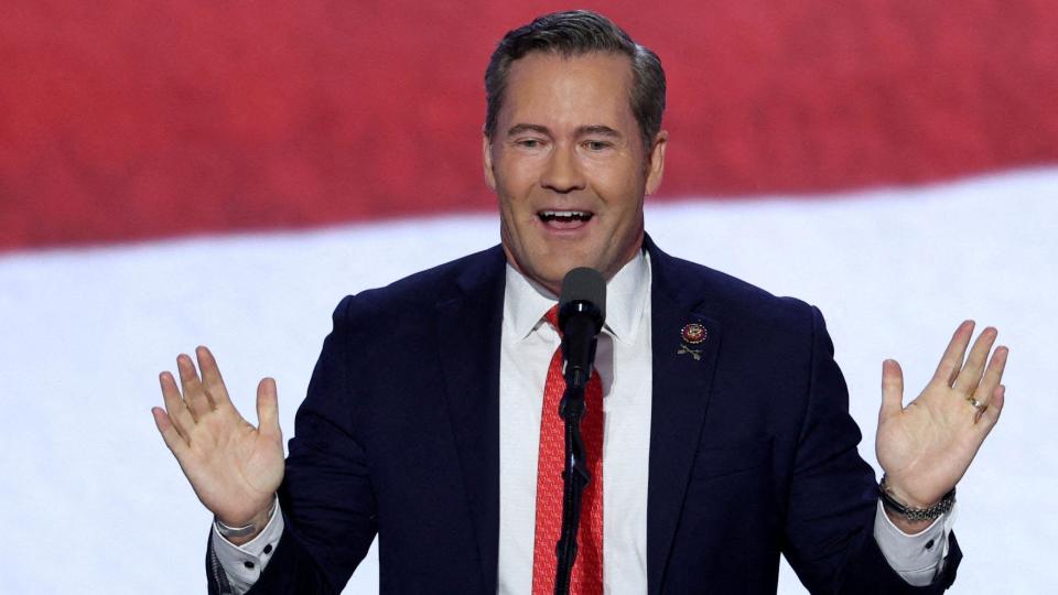 Rep. Mike Waltz stands in front of a red and white striped background wearing a blue blazer, white shirt and red tie. He is smiling and holding his hands up while speaking into a microphone