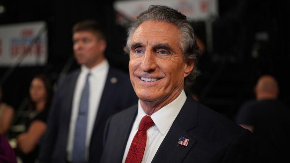 Doug Burgum, with salt and pepper hair, and wearing a black suit with a red tie, smiles for the cameras