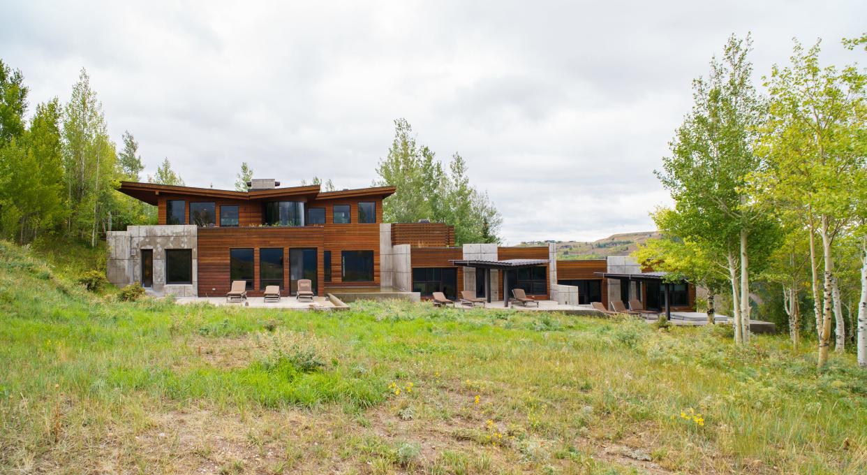 The exterior of a large, long house made of wood and concrete with an outdoor patio and nine lawn chairs in a green field with trees on the left and right with mountains in the background