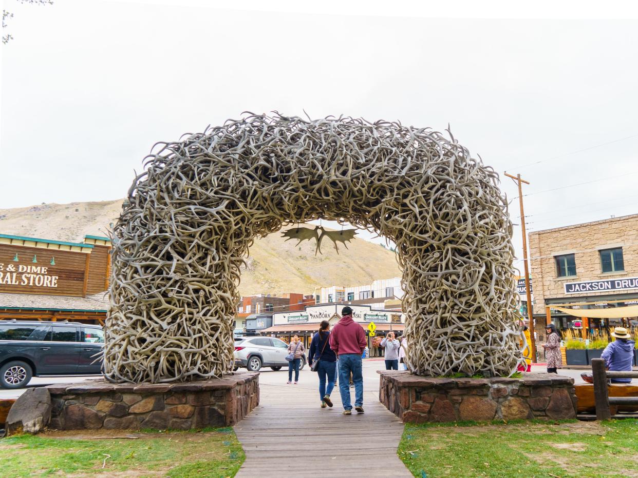 People walk through an arch made of antlers on a busy street lined with shops in the background