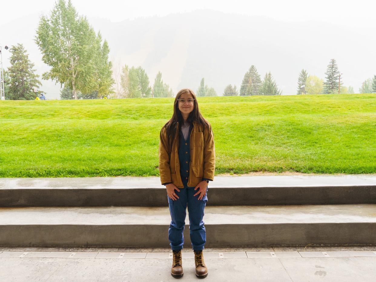 The author dressed in layers stands in front of a hazy scene with green fields and trees in the background