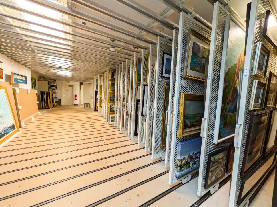 A photo of an organized art storage room featuring sliding metal racks filled with framed paintings of various styles and sizes. The space is well-lit with overhead lighting, and the racks are neatly arranged along tracks on the floor, showcasing an efficient system for storing and displaying artwork.