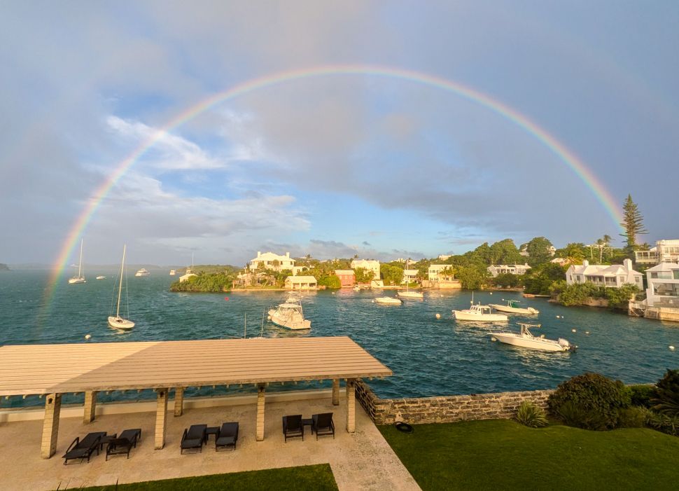 A photo of a vibrant rainbow stretching across a picturesque harbor in Bermuda, with boats floating on the water and whitewashed houses dotting the shoreline under a partly cloudy sky.