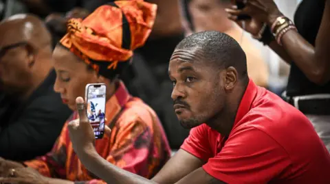 PHILIPPE LOPEZ/AFP The leader of the Rally for the Protection of Afro-Caribbean Peoples and Resources (RPPRAC) Rodrigue Petito, wearing a red T-shirt, looks at his phone, during what appears to be a meeting. 