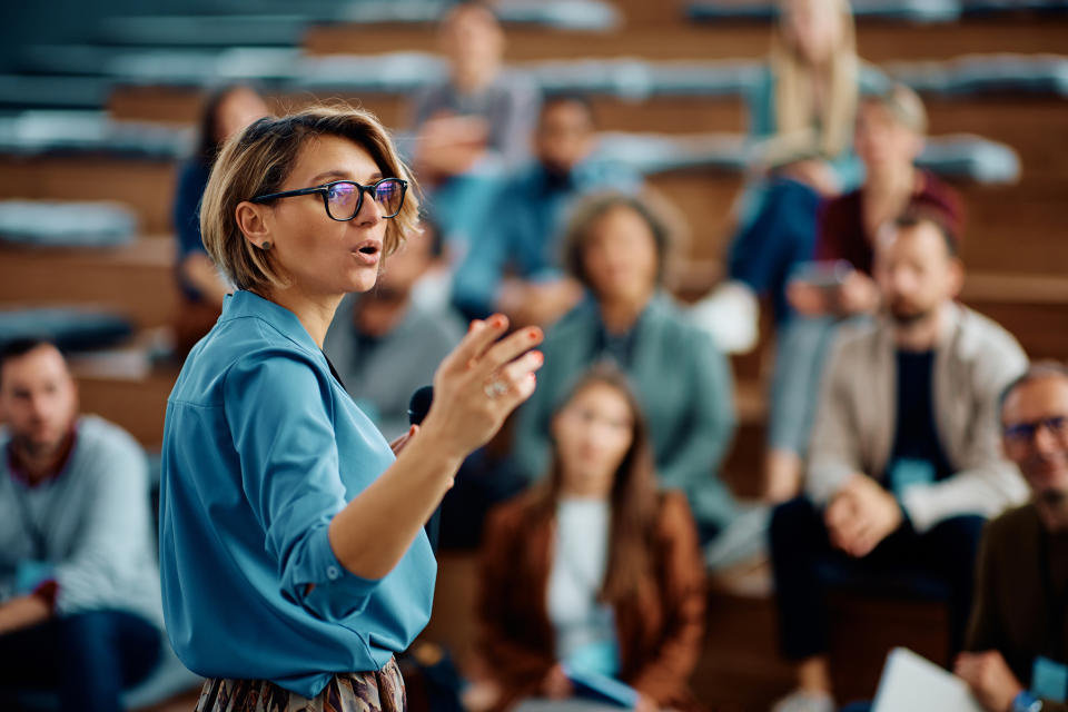 A person in glasses and a blue shirt speaks to a seated audience in an auditorium