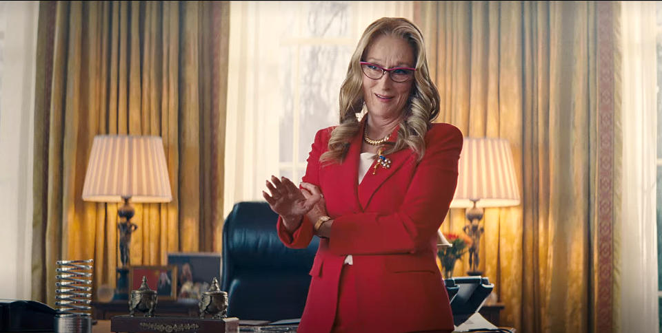 Person in an office wearing a tailored suit and glasses, gesturing with hands