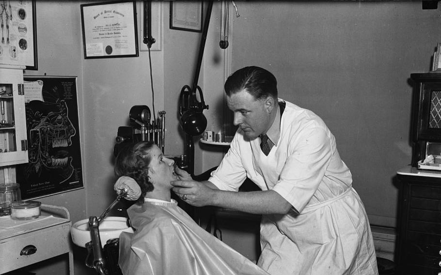 A dentist in a white coat looks into a woman’s mouth as she’s seated on a chair.