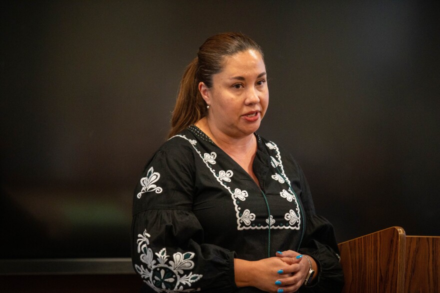 A woman in a black and white embroidered shirt crosses her blue painted nails in front of her as she speaks standing next to a podium 