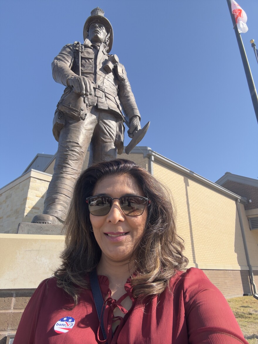 A woman wearing a maroon top and sunglasses stands in front of the statue of a fire fighter at the Frisco Fire Station, which served as a polling location during early voting.