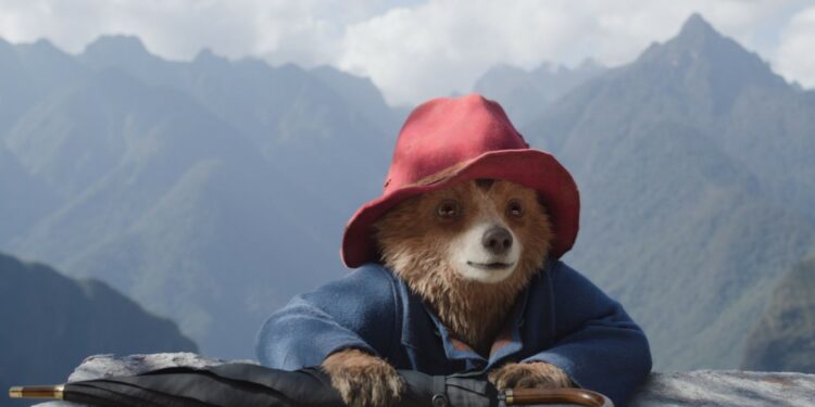 A bear wearing a red hat looks over a rock, with mountains behind.