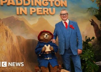 Kerry Evans posing for a photo in Gadebridge Pharmacy in Hemel. She is wearing her uniform - a royal blue NHS coloured shirt with a collar with white edging and black cardigan. She is holding a jar of "Aunt Lucy's Marmalade" which has a Peruvian cloth on the top. She has highlighted blonde hair partly swept back into a clip and behind her are shelves of chemist goods.