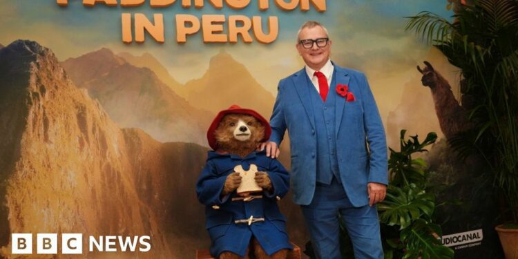 Kerry Evans posing for a photo in Gadebridge Pharmacy in Hemel. She is wearing her uniform - a royal blue NHS coloured shirt with a collar with white edging and black cardigan. She is holding a jar of "Aunt Lucy's Marmalade" which has a Peruvian cloth on the top. She has highlighted blonde hair partly swept back into a clip and behind her are shelves of chemist goods.