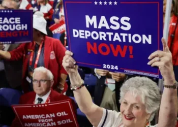 An elderly Trump supporter in a white and red outfit holds a card that reads