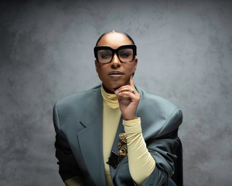 A Black woman wearing a green blazer and pale yellow turtleneck with her hair pulled back and large square black glasses looks directly at the camera.  