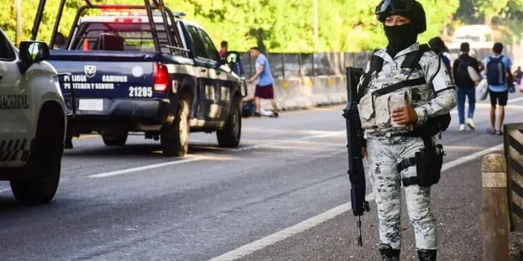 National Guardsperson in full uniform and carrying an automatic gun stands at duty on a street in Tapachula, Chiapas, filled with state police and national guard trucks