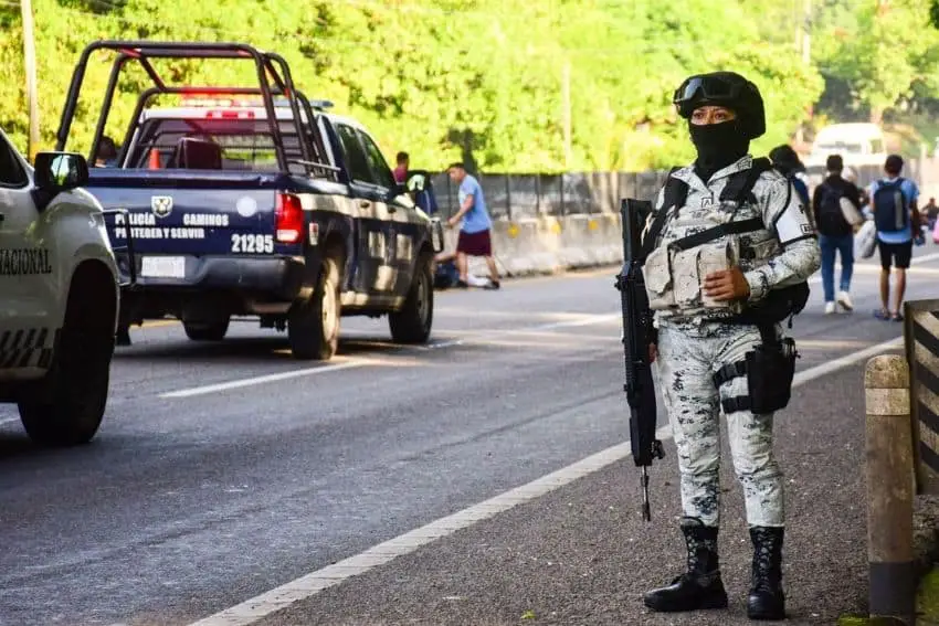 National Guardsperson in full uniform and carrying an automatic gun stands at duty on a street in Tapachula, Chiapas, filled with state police and national guard trucks