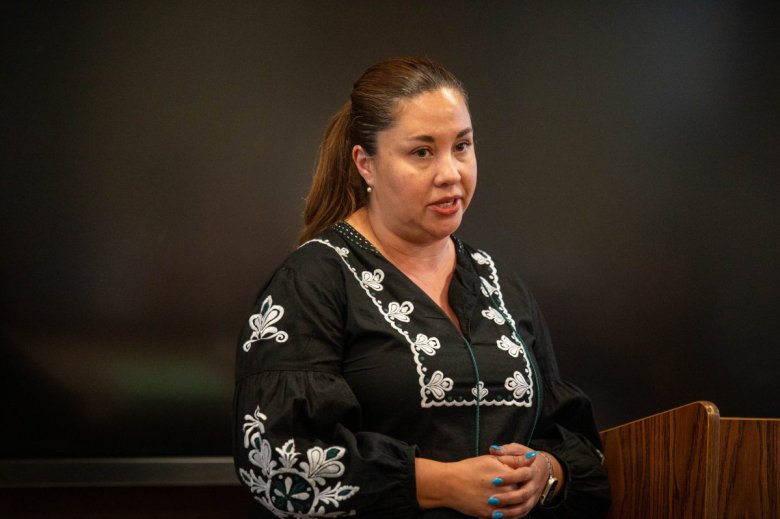 Yadira Caraveo in a black embroidered blouse stands at a podium, speaking in front of a dark background.