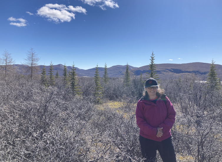 A woman standing among trees and shrubs with mountain landscape behind her.