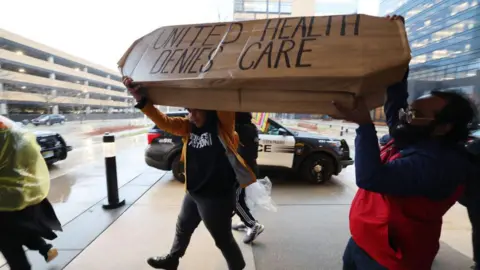 Getty Images Protesters carry a fake coffin that reads: United Health Denies Care